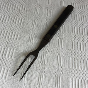 Peut inclure: Une fourchette à découper vintage avec un manche en bois foncé et deux dents. La poignée est fixée à la fourchette avec des rivets. La fourchette est en métal et présente des signes d'âge et d'utilisation. Le fond est une surface blanche texturée.
