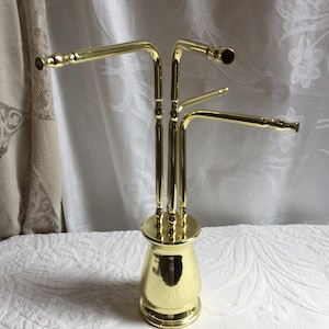 Puede incluir: Un toallero de metal dorado con múltiples brazos curvos que se extienden desde una base central. El toallero tiene un diseño decorativo y ornamentado, y probablemente se utiliza para colgar toallas en un baño.