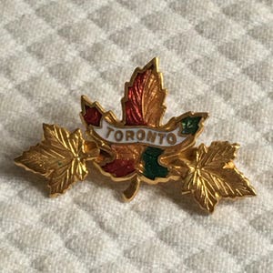 Op de afbeelding: Een goudkleurige esdoornbladspeld met het woord "TORONTO" op een witte banner. Het esdoornblad heeft rode, groene en bruine accenten. Twee kleinere gouden bladeren bevinden zich aan weerszijden.