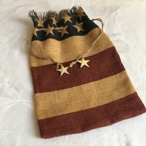 May include: A burlap drawstring bag with a red and tan striped pattern and black stars. The bag has a frayed edge and is decorated with three gold stars.