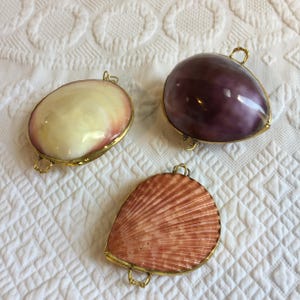 May include: Three decorative shell pendants with gold-toned metal edges and small loops. One shell is smooth and cream-colored, one is a polished deep purple, and the third is a textured, ribbed shell in a soft peach-orange hue.