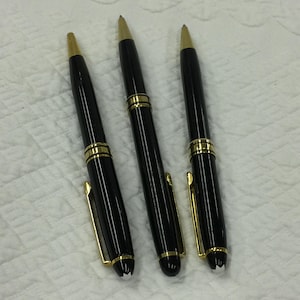 May include: Three black pens with gold-coloured accents. Each pen has a gold-coloured tip, a gold band near the centre, and a gold clip. The pens are arranged on a white, textured surface.