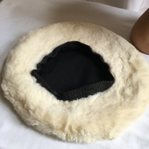 Puede incluir: Una boina cremosa y esponjosa con interior negro y ribete gris oscuro. La boina tiene un aspecto suave y texturizado, lo que sugiere un accesorio cálido y cómodo. El sombrero es de forma circular.