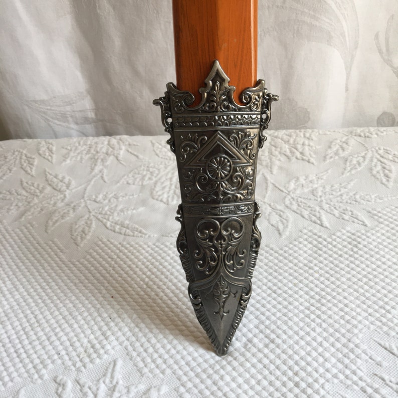 Puede incluir: Una vaina de espada de color plateado con un dise&ntilde;o ornamentado. La vaina es puntiaguda en la parte inferior y tiene una parte superior decorativa.