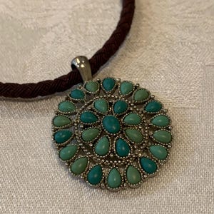 Puede incluir: Un collar con colgante plateado con un diseño circular de piedras turquesas. El colgante presenta un grupo de piedras turquesas en forma de lágrima. El collar tiene un cordón trenzado marrón oscuro.