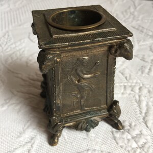 May include: A small, square, bronze inkwell with a decorative design featuring a figure in relief. The inkwell has a round opening at the top and sits on four feet.