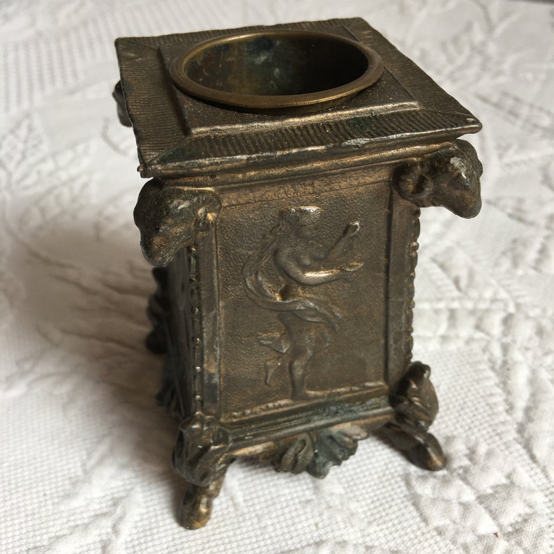 May include: A small, square, bronze inkwell with a decorative design featuring a figure in relief. The inkwell has a round opening at the top and sits on four feet.