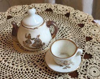 May include: A miniature porcelain tea set with a white teapot and cup and saucer. The teapot has a gold trim and a design of two women in silhouette. The cup and saucer also have gold trim and a floral design.