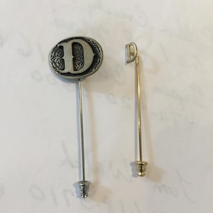 May include: Two stick pins. One has a silver-coloured, oval-shaped head with the letter "D" in the centre, surrounded by a decorative border. The other has a gold-coloured head with the letter "D".