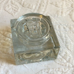 Puede incluir: Un tintero de vidrio transparente con una base cuadrada y una tapa redonda. La tapa tiene un diseño en relieve de dos figuras que se toman de la mano.