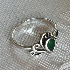 May include: A silver ring with a teardrop-shaped, emerald-green stone. The ring features ornate scrollwork around the stone and along the band. The ring is set against a light gray fabric background.