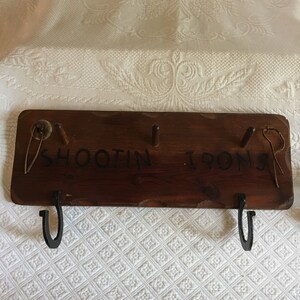 May include: A wooden sign with the words "Shootin Irons" painted on it. The sign has three hooks on the bottom and a small metal ring on the right side.