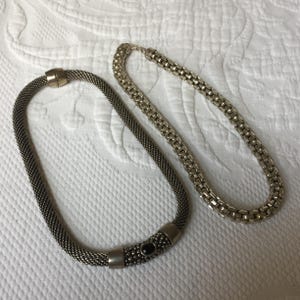 May include: Two silver necklaces, one with a black stone accent. The necklace on the left has a more intricate design and a black stone accent. The necklace on the right has a more simple design.