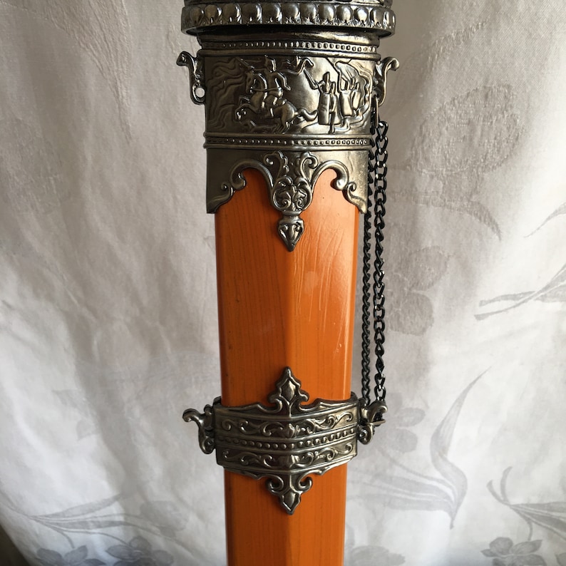Puede incluir: Primer plano de la parte superior de una vaina de espada decorativa. La vaina est&aacute; hecha de madera y pintada de naranja. La parte superior de la vaina est&aacute; decorada con metal plateado y una cadena.