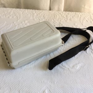 May include: A white rectangular case with a black strap. The case has a textured surface and a zipper closure.
