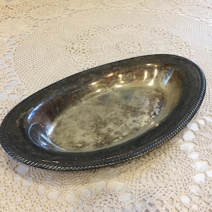 May include: An oval silver-plated serving dish with a rope-style edge. The interior of the dish shows signs of wear and age, with a tarnished appearance. The dish is empty and rests on a white lace tablecloth.