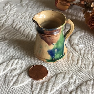 Könnte beinhalten: Ein kleiner, handbemalter Keramik Krug mit brauner, grüner und blauer Glasur. Der Krug hat einen runden Körper und einen kleinen Henkel. Er steht auf einer weißen Oberfläche.