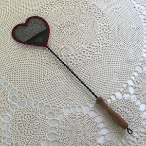 Puede incluir: Una matamoscas en forma de corazón con una cabeza de malla oscura y un borde burdeos. El mango es de madera y el tallo largo y delgado es negro. El matamoscas está sobre un fondo crema tejido a ganchillo.
