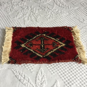 May include: A rectangular woven rug featuring a deep red background with a central diamond pattern in black and cream. The rug has a thick, textured pile and is finished with cream-colored fringe tassels on the two shorter opposing ends.