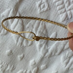 May include: A gold-colored bracelet with a delicate, woven design. The bracelet features a hook-and-eye clasp. The jewelry is displayed on a white, textured surface. This bracelet is a piece of fashion jewelry.