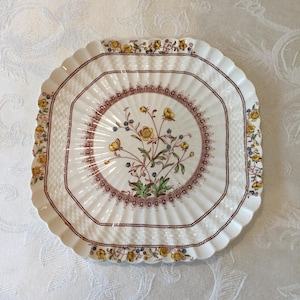 Puede incluir: Un plato decorativo blanco, de forma cuadrada, con borde festoneado. El plato presenta un diseño floral con flores amarillas, hojas verdes y bayas azules. Un borde marrón rodea el diseño central y el borde del plato.