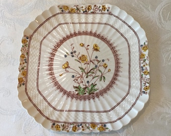 Op de afbeelding: Een witte, vierkante decoratieve plaat met een geschulpte rand. De plaat heeft een bloemenontwerp met gele bloemen, groene bladeren en blauwe bessen. Een bruine rand omringt het centrale ontwerp en de rand van de plaat.