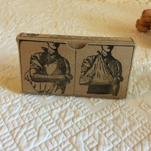 Puede incluir: Una caja de cartón vintage con instrucciones sobre cómo usar una bandolera. La caja es marrón y tiene texto negro que dice "May Be Used To Follow Instructions Distributed by MEDICAL SUPPLY CO., Rockford, Ill.". La caja muestra dos ilustraciones de una persona con una bandolera en el brazo.
