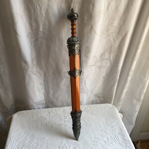 Puede incluir: Una espada decorativa con mango de madera y detalles en metal plateado. La espada tiene una vaina de metal plateado con dise&ntilde;os intrincados.