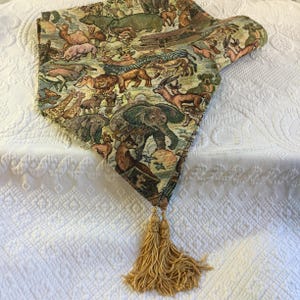 Puede incluir: Camino de mesa decorativo con un diseño de tapiz que representa varios animales, como elefantes, jirafas y cebras. El camino de mesa tiene una borla beige y está hecho de tela tejida.