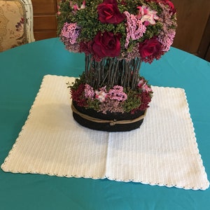 May include: A white crocheted table runner with a scalloped edge sits on a teal tablecloth. A floral arrangement with red and pink flowers is in a brown woven basket on top of the runner.