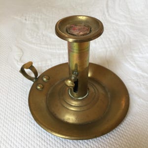Puede incluir: Un candelero de latón con una base redonda y un portavelas extraíble. El candelero tiene una pequeña manija en el lateral y una perilla en la parte delantera.