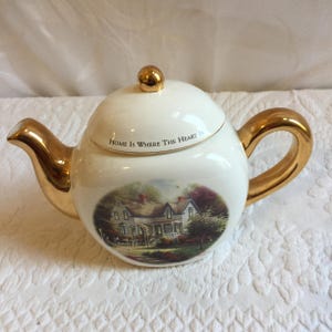 May include: White teapot with a gold handle and trim. The teapot has a picture of a house with a tree and the words "Home is where the heart is" on the side.