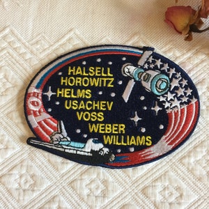 May include: A blue and red embroidered patch featuring a space shuttle and the names of the crew members: Halsell, Horowitz, Helms, Usachev, Voss, Weber, and Williams.