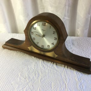 May include: A wooden mantel clock with a rounded top and a wide base. The clock face is silver with black Arabic numerals and the text Gilbert. It features a gold-colored metal bezel surrounding the dial and a gold-colored accent plate below.