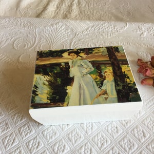 Puede incluir: Caja de madera blanca con una imagen pintada de dos mujeres en un entorno de parque. Las mujeres llevan vestidos largos y sombreros. El fondo es un paisaje verde y azul con un estanque.