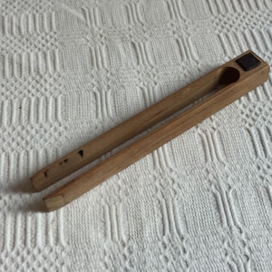 May include: A wooden clothespin with a rectangular block at the top and a small, dark square attached. The clothespin is light brown and has a simple design. The two arms of the clothespin are parallel and taper to a point.