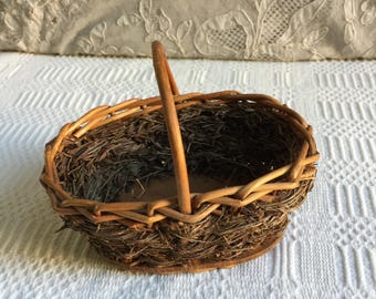 Puede incluir: Cesta ovalada tejida con asa de madera. La cesta está hecha de ramitas marrones y tiene un aspecto rústico. El interior está lleno de hierba seca. La cesta se encuentra sobre una superficie blanca.