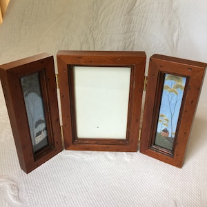 May include: A tri-fold wooden picture frame with a center opening and two smaller side openings. Each opening features a painted scene of a house with trees in a winter landscape.