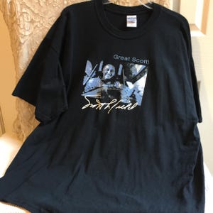 Puede incluir: Camiseta negra con un gráfico de un hombre y el texto "Great Scott!" y una firma. El gráfico tiene acentos azules y blancos. La camiseta está hecha de un material suave.