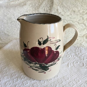 May include: A beige ceramic pitcher with a brown rim and a handle. The pitcher has a painted design of three red apples with green leaves on a white background.