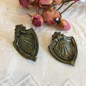 May include: Two antique bronze-colored shield-shaped drawer pulls with a weathered, aged appearance. Each pull features a decorative shell design within the shield and ornate detailing at the top. The pulls are set against a white textured background.