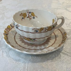 Puede incluir: Juego de taza y platillo de té blanco con diseño floral. La taza y el platillo tienen un borde festoneado y un diseño de cesta tejida. El diseño floral incluye flores amarillas y azules con hojas verdes. El juego es de cerámica.
