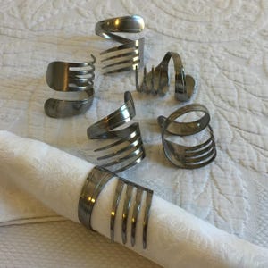 Puede incluir: Seis anillos de servilleta de plata hechos de cubiertos doblados. Los anillos están dispuestos sobre una superficie blanca.