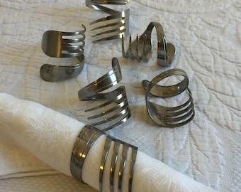 May include: Six silver fork napkin rings made from bent silverware. The rings are arranged on a white surface.