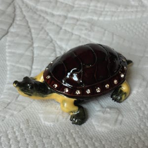 May include: A small, dark brown turtle figurine with a yellow underside. The shell is decorated with a row of clear rhinestones.