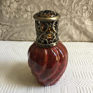 Puede incluir: Lámpara de aceite decorativa con base de cerámica vidriada roja y marrón. La lámpara presenta una parte superior de metal de filigrana de color bronce. La base tiene un diseño texturizado y arremolinado. Para la decoración del hogar.