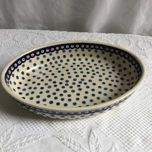 May include: An oval ceramic bowl with a cream-colored base featuring a pattern of small dark blue dots. The rim is decorated with a repeating border of dark blue circles with orange centers, creating a consistent design around the entire edge.