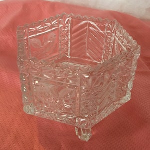 May include: A clear glass bowl with a geometric design and three legs. The bowl has a square shape and is decorated with intricate patterns.