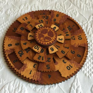 May include: A circular wooden puzzle featuring multiple rotating gear-shaped layers with serrated edges. The surface is stained in shades of brown and tan, displaying black printed numbers and cross-hatched patterns across the interlocking segments.