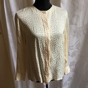 Puede incluir: Una blusa de manga larga de color crema con un estampado animal sutil de color marrón claro. La blusa tiene un cierre de botones en la parte delantera y un ajuste holgado.